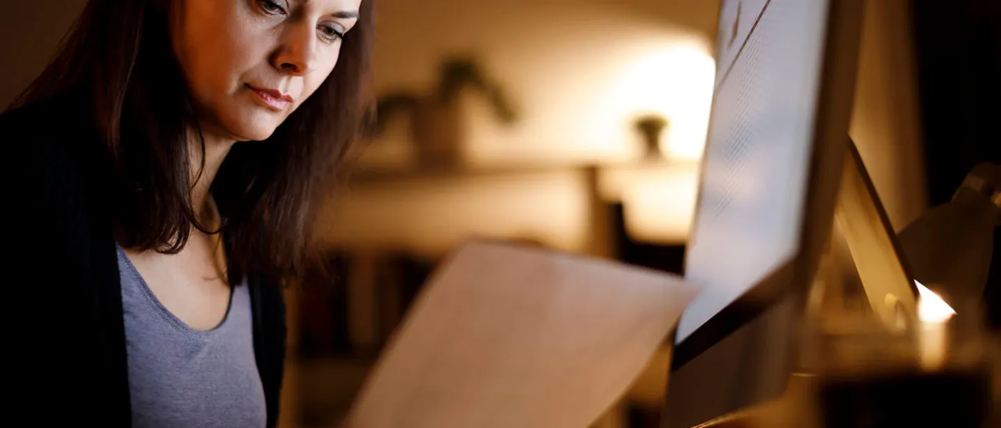 Una mujer sentada leyendo una hoja de papel frente a la pantalla de un computador